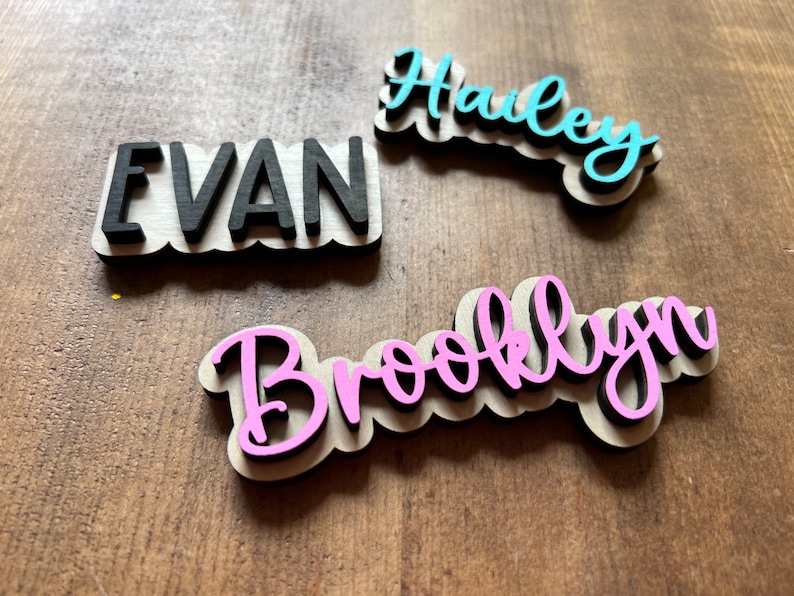 May include: Three wooden name plaques with names "Evan", "Hailey", and "Brooklyn" written in black, blue, and pink paint, respectively. The plaques are on a wooden surface.