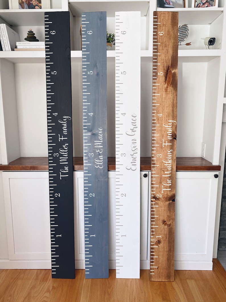 May include: Four wooden growth charts with white painted markings and names. The charts are painted in black, blue, white, and brown. The names on the charts are "The Miller Family", "Ella & Maeve", "Emerson Grace", and "The Watkins Family".