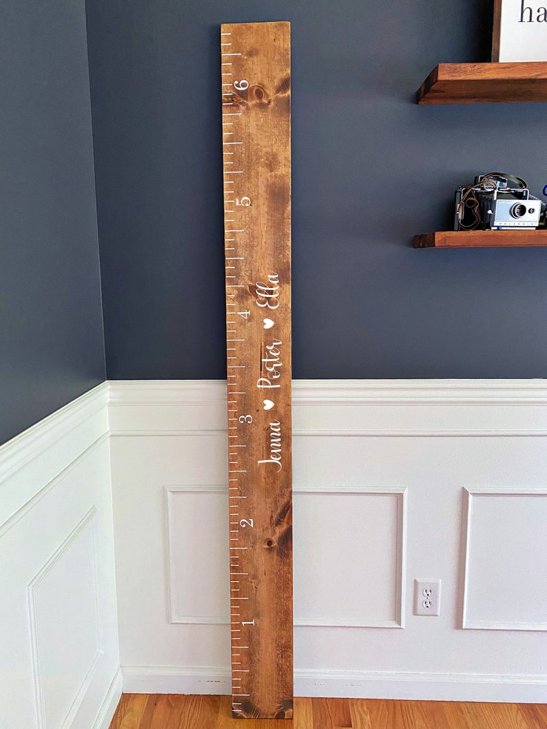 May include: A wooden growth chart with a ruler marked in inches from 1 to 6. The chart is stained a dark brown color and has white text that reads "Jenna & Peter Ella" with a heart symbol between each name.