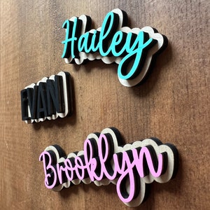 May include: Three wooden name signs with black outlines. The names are "Haley" in turquoise, "Evan" in black, and "Brooklyn" in pink.