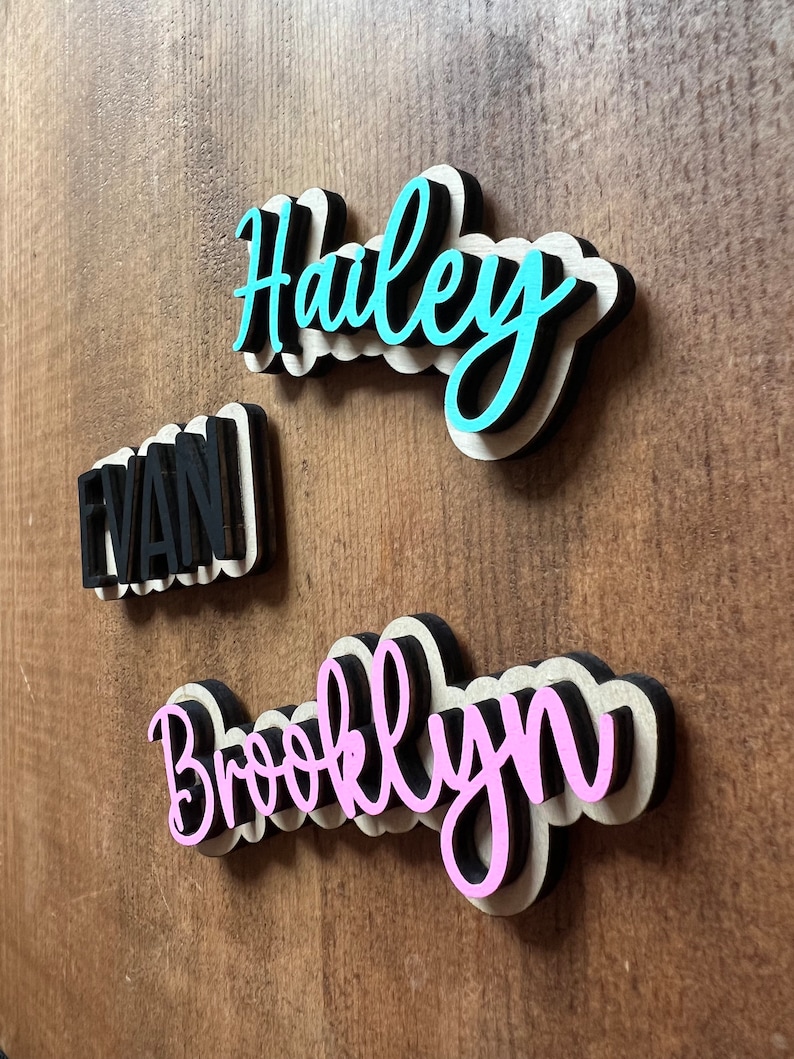 May include: Three wooden name signs with black outlines. The names are "Haley" in turquoise, "Evan" in black, and "Brooklyn" in pink.