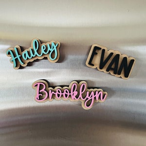 May include: Three wooden name plaques with the names "Hailey", "Evan", and "Brooklyn" written in cursive. The names are painted in different colors: Hailey is turquoise, Evan is black, and Brooklyn is pink.