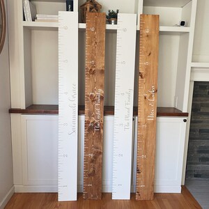 May include: Four wooden growth charts with white lettering. The charts are mounted on a white wall with a wooden shelf above them. The charts are labeled "Savannah Grace", "The Casti Family", "The Mead Family", and "Alice & Luka".
