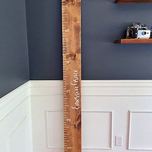 May include: A wooden growth chart with a ruler marked in inches from 1 to 6. The chart is stained a dark brown color and has the name "Emerson Grace" written in white lettering.