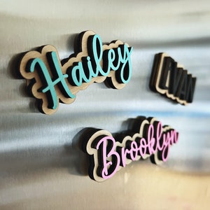 May include: Three wooden name magnets with black outlines and colorful lettering. The names are Hailey in teal, Brooklyn in pink, and Evan in black.