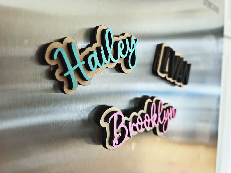 May include: Three wooden name magnets with black outlines and colorful lettering. The names are Hailey in teal, Brooklyn in pink, and Evan in black.