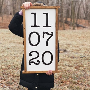 K&ouml;nnte beinhalten: Ein wei&szlig;es Holzschild mit braunem Rahmen, das schwarze Zahlen mit der Aufschrift "11 07 20" zeigt.
