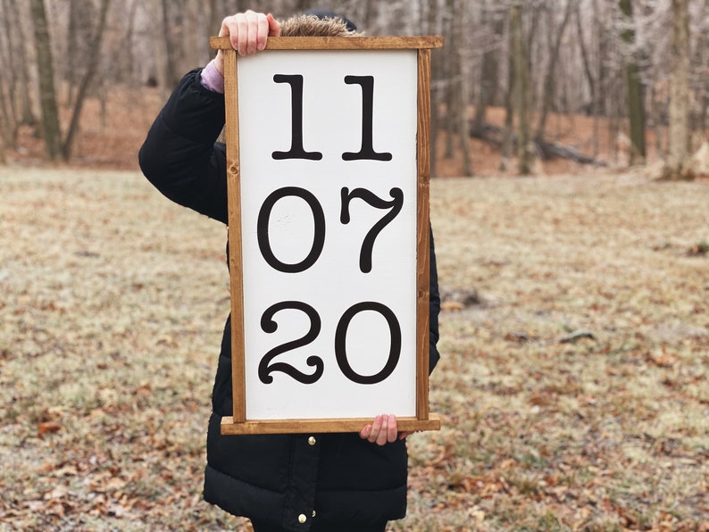 K&ouml;nnte beinhalten: Ein wei&szlig;es Holzschild mit braunem Rahmen, das schwarze Zahlen mit der Aufschrift "11 07 20" zeigt.