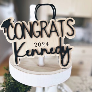 May include: Wooden graduation ornament with black lettering that reads "Congrats Kennedy 2024". The ornament features a graduation cap icon.