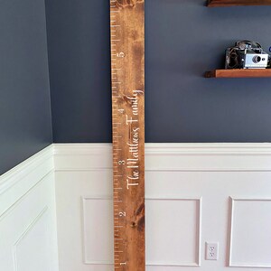 May include: A brown wooden growth chart with white numbers and the text "The Matthews Family" printed on it. The chart is mounted on a wall with white wainscoting.