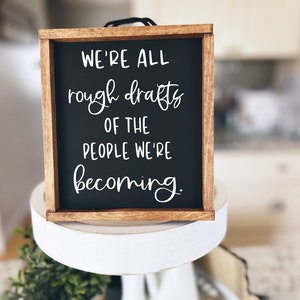 May include: A wooden framed sign with a black background and white text that reads "We're all rough drafts of the people we're becoming."
