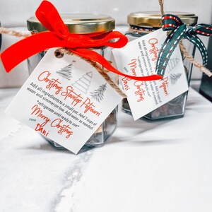 May include: Three glass jars with gold lids and red, green, and white ribbon tied around the lids. Each jar contains a mixture of dried herbs and spices. The jars are labelled with white paper tags that say "Christmas Stovetop Potpourri" and include instructions for use. The tags are decorated with a Christmas tree design.