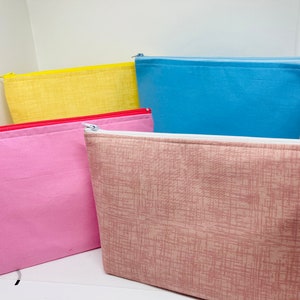 May include: Four zippered pouches in different colours and patterns. The pouches are made of fabric and have a zipper closure. The pouches are stacked on top of each other. The top pouch is light blue, the second pouch is yellow, the third pouch is pink, and the bottom pouch is a light brown with a subtle pattern.