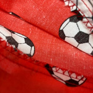 Op de afbeelding: Rode stof met een patroon van witte en zwarte voetballen. De stof is dubbelgevouwen en de naden zijn zichtbaar.