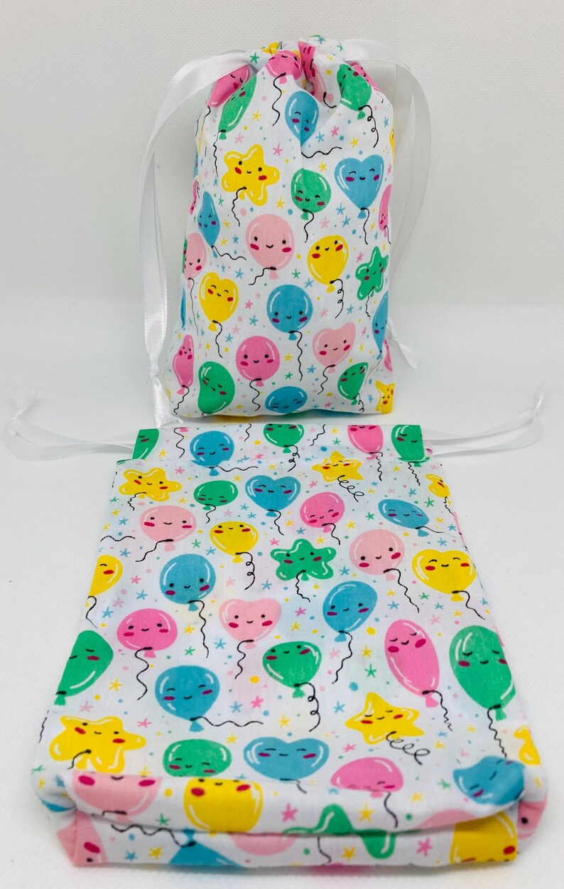 Op de afbeelding: Twee witte trekkoordtassen met een kleurrijk patroon van ballonnen en sterren. De ballonnen hebben vrolijke gezichten en zijn in tinten roze, geel, blauw en groen.