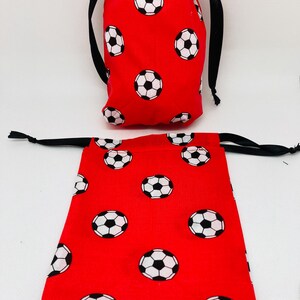 Op de afbeelding: Twee rode trekkoordtassen met een wit voetbalpatroon. De kleinere tas staat rechtop, terwijl de grotere tas plat ligt.