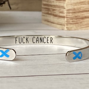 May include: A silver-colored cuff bracelet with the words "FUCK CANCER" engraved on the outside. The bracelet has a blue ribbon design on each end, symbolizing cancer awareness.
