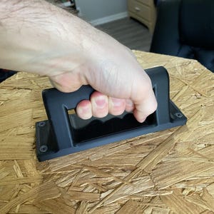 May include: A black plastic handle with a rectangular opening is attached to a piece of plywood.