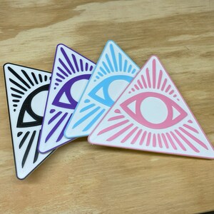 May include: Four triangle-shaped stickers with a white eye design inside. The stickers are in black, purple, blue, and pink.