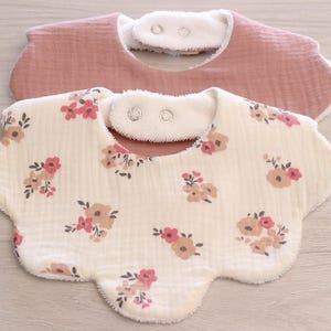 Peut inclure: Deux bavoirs à bords festonnés avec fermetures à pression. Le bavoir supérieur est rose poudré uni, tandis que le bavoir inférieur est crème avec un motif floral répétitif dans des tons de rose, beige et vert foncé. Tissu texturé doux.