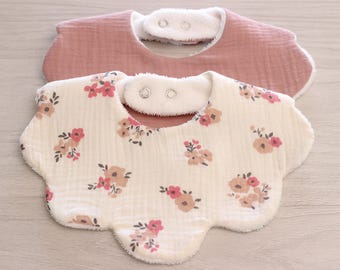 May include: Two scalloped-edge bibs with snap closures. The top bib is solid dusty rose, while the bottom bib is cream-colored with a repeating floral pattern in shades of pink, tan, and dark green. Both feature a soft, textured fabric finish.