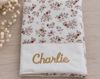May include: A rectangular fabric cover featuring a white background with a repeating floral pattern in shades of brown, rust, and beige. The lower section is a solid white band with the name Charlie embroidered in a gold-colored script font.