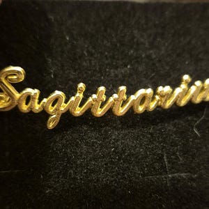 May include: A gold-coloured bracelet featuring the word "Sagittarius" in a cursive script. The bracelet is set against a dark backdrop. The jewellery is likely made of gold or a gold-coloured metal.