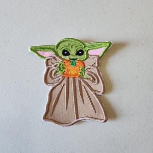 May include: Green and brown embroidered patch of a character holding an orange pumpkin. The character has large eyes and a small mouth.