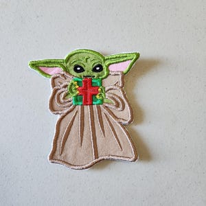 May include: Green and brown embroidered patch of a character holding a green and red gift box. The character has large eyes and a pink collar.