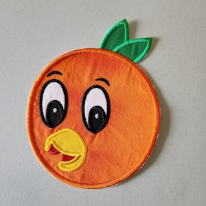 Könnte beinhalten: Orangefarbener Filzapplikation einer Cartoon-Orange mit einem lächelnden Gesicht, zwei großen Augen und einem gelben Schnabel. Die Orange hat zwei grüne Blätter oben.