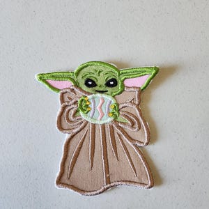 May include: Embroidered patch featuring a green alien character with large ears and black eyes, holding a pastel-colored Easter egg. The character is wearing a tan robe with a white border. The patch is designed for application on fabric.