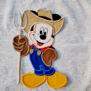 Può includere: Un personaggio dei cartoni animati che indossa un cappello da cowboy e una salopette, tenendo un forcone. Il personaggio è marrone, giallo, blu e rosso.