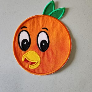 Könnte beinhalten: Ein runder orangefarbener Filz-Patch mit einem Comic-Gesicht, großen Augen und einem gelben Schnabel. Die Orange hat ein grünes Blatt oben drauf.