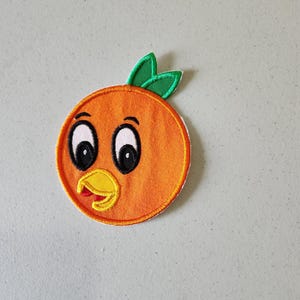 Könnte beinhalten: Orangefarbener gestickter Aufnäher mit einem Comic-Gesicht, zwei großen Augen, einem gelben Schnabel und grünen Blättern oben.