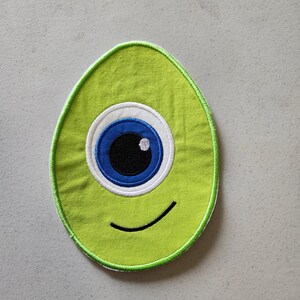 May include: Green felt patch with a single blue eye and a smiling mouth. The eye has a white outline and a black pupil.