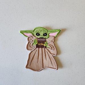 May include: Green and brown embroidered patch of a character from the Star Wars franchise, holding a book with the word "Baby" on it.