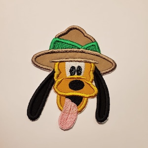 Könnte beinhalten: Ein gestickter Aufnäher von Pluto dem Hund, der einen braunen und grünen Rangerhut trägt. Pluto streckt seine Zunge heraus.