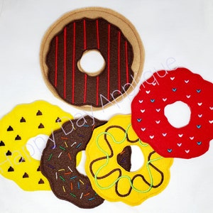 Könnte beinhalten: Fünf Filzdonuts in verschiedenen Farben und Designs. Ein brauner Donut mit roten Streifen, ein roter Donut mit weißen Herzen, ein gelber Donut mit braunen Schokoladenstückchen, ein brauner Donut mit bunten Streuseln und ein gelber Donut mit einem grünen Wirbel und brauner Mitte.