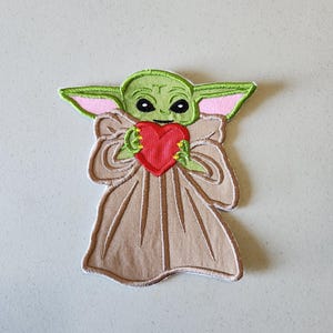 May include: Green and brown embroidered patch of a character from the Star Wars franchise, holding a red heart.