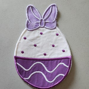 May include: Purple and white Easter egg shaped patch with a white bow on top. The egg has white polka dots and a wavy purple and white design at the bottom.