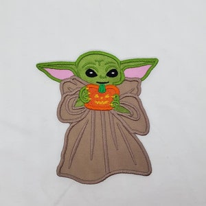 May include: Green and brown embroidered patch of a character holding a jack-o'-lantern. The character has large ears and big eyes.