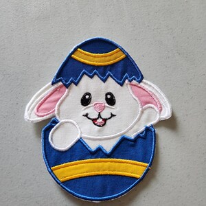 May include: A white bunny rabbit peeking out of a blue and yellow Easter egg. The egg is cracked open and the bunny is smiling.