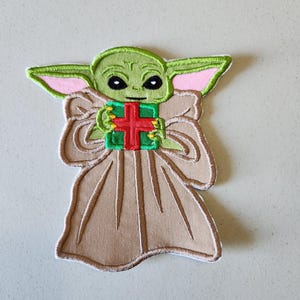 May include: Green and brown embroidered patch of a character holding a red and green gift box. The character has large ears and a big smile.