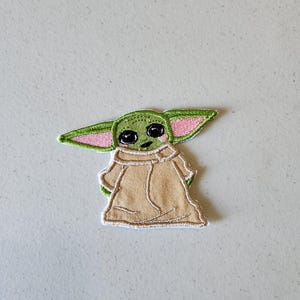 Peut inclure: Patch brodé vert et rose d'un personnage de la franchise Star Wars, connu sous le nom de "L'Enfant" ou "Baby Yoda", portant une robe brune.