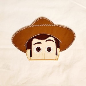 May include: Embroidered patch of Woody from Toy Story. The patch features Woody's face with his signature brown hat and a light brown rope trim.
