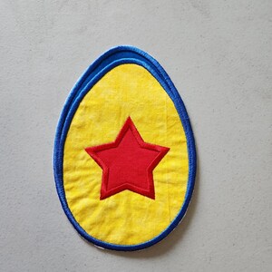 May include: A yellow felt egg with a red star in the centre, surrounded by a blue felt border.