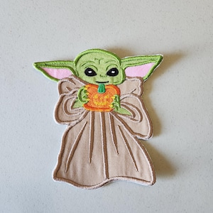 May include: Green and brown embroidered patch of a character holding a pumpkin. The character has large eyes and a small mouth. The pumpkin is orange with a green stem.