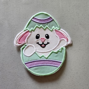 May include: A green and purple Easter egg with a white bunny peeking out of the top. The bunny has pink ears and a pink nose.