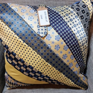 May include: A square throw cushion constructed from diagonal strips of various neckties in shades of navy blue, gold, and cream with diverse geometric and floral patterns. A small brown paper tag is attached, featuring the text With lots of Love.