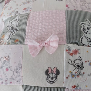 以下が含まれることがあります： ピンク、グレー、白の色合いのパッチワーク枕。バンビとミニーマウスの刺繍、花柄、ピンクのリボンが特徴です。枕はコーデュロイの質感です。寸法はセンチメートル単位です。